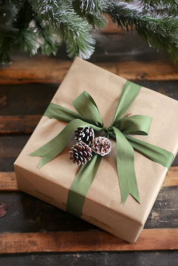 Gift wrapped in brown paper with a green ribbon and pine cones on a wooden surface.